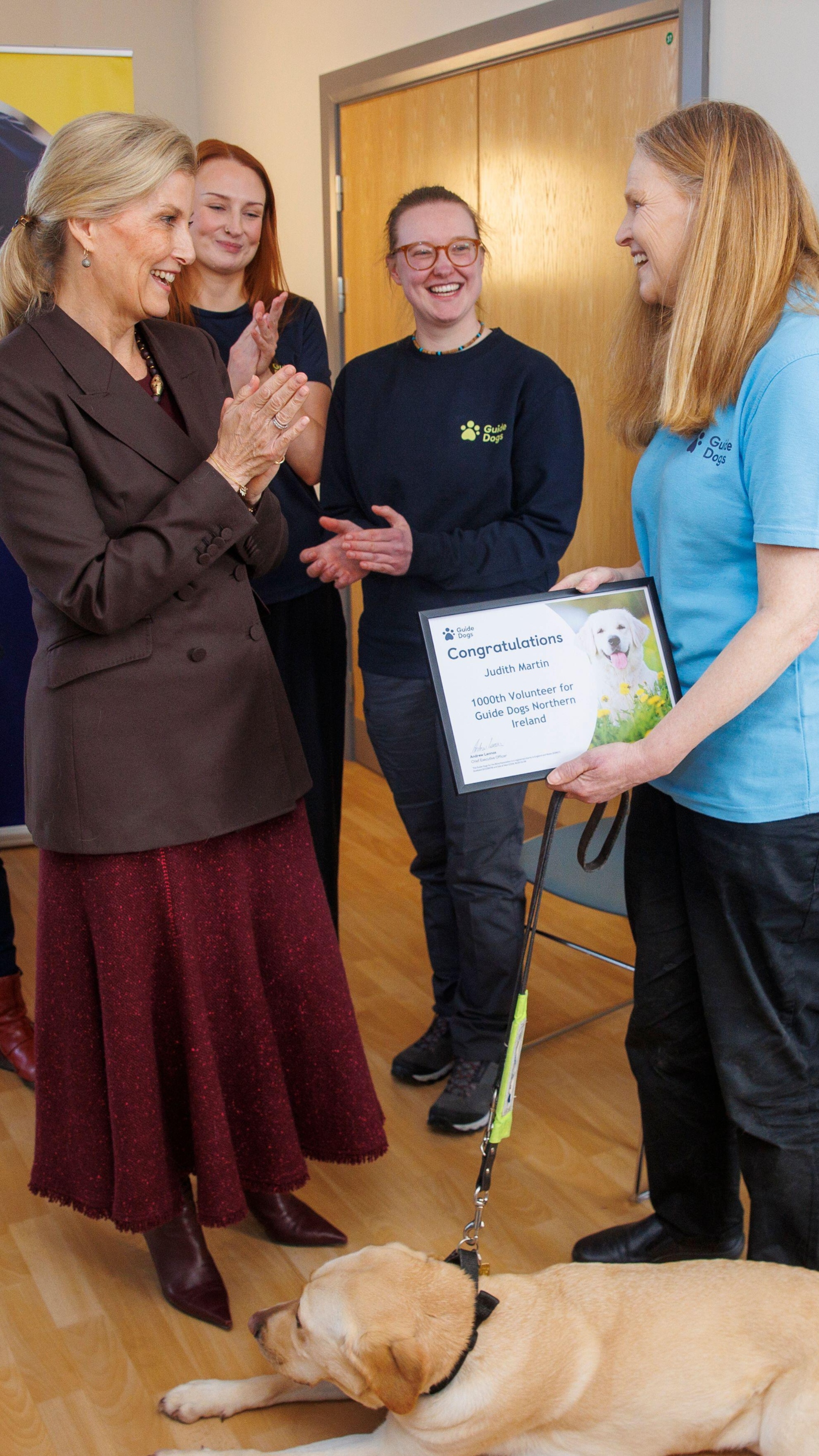 The Duchess of Edinburgh (centre left) presents Judith Martin (right) and her guide dog Brooke with a certificate for being the 1000th active volunteer for Guide Dogs Northern Ireland