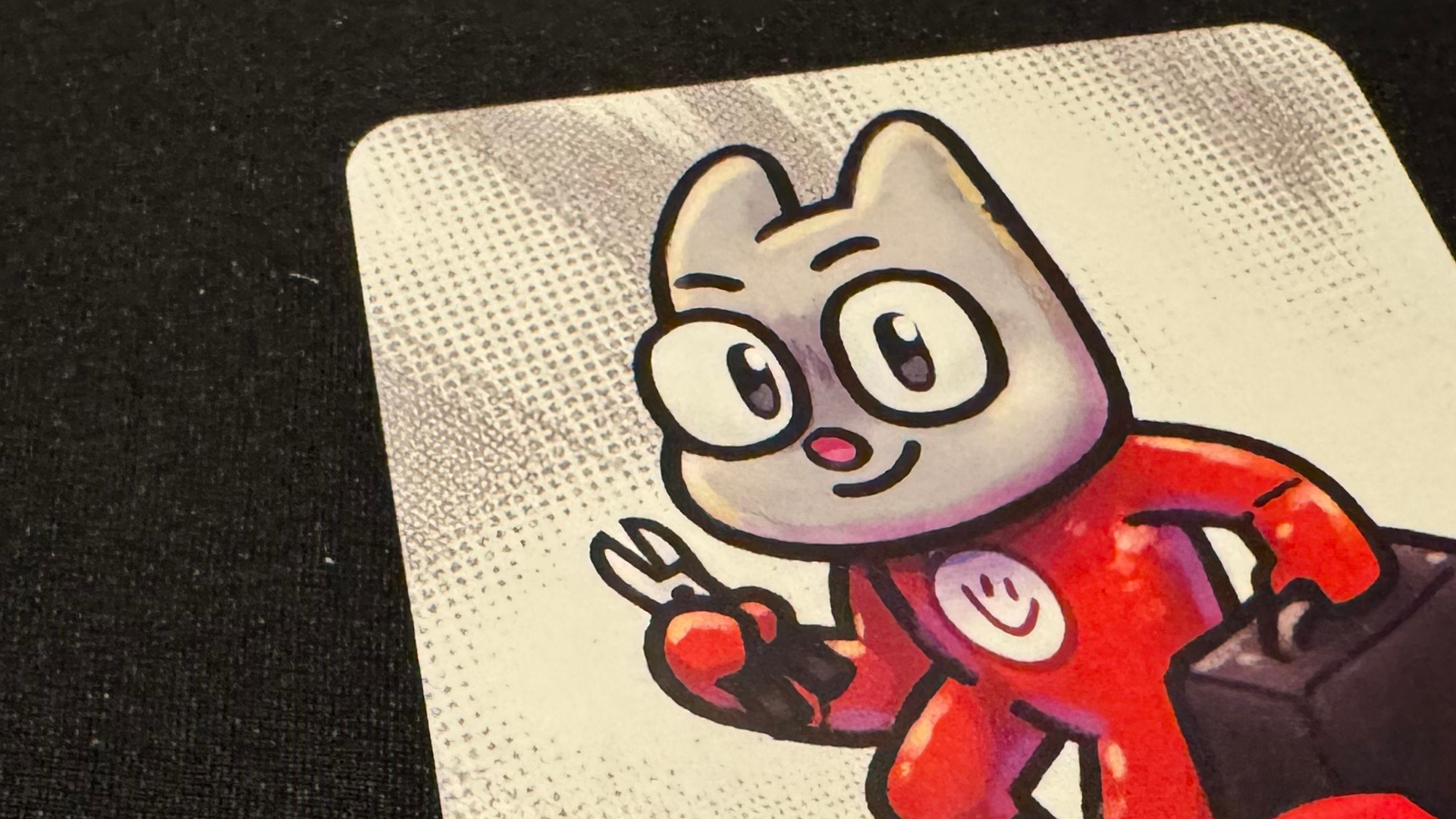 A cat-esque creature dressed in red overalls holding clippers on a card, which sits on a black surface