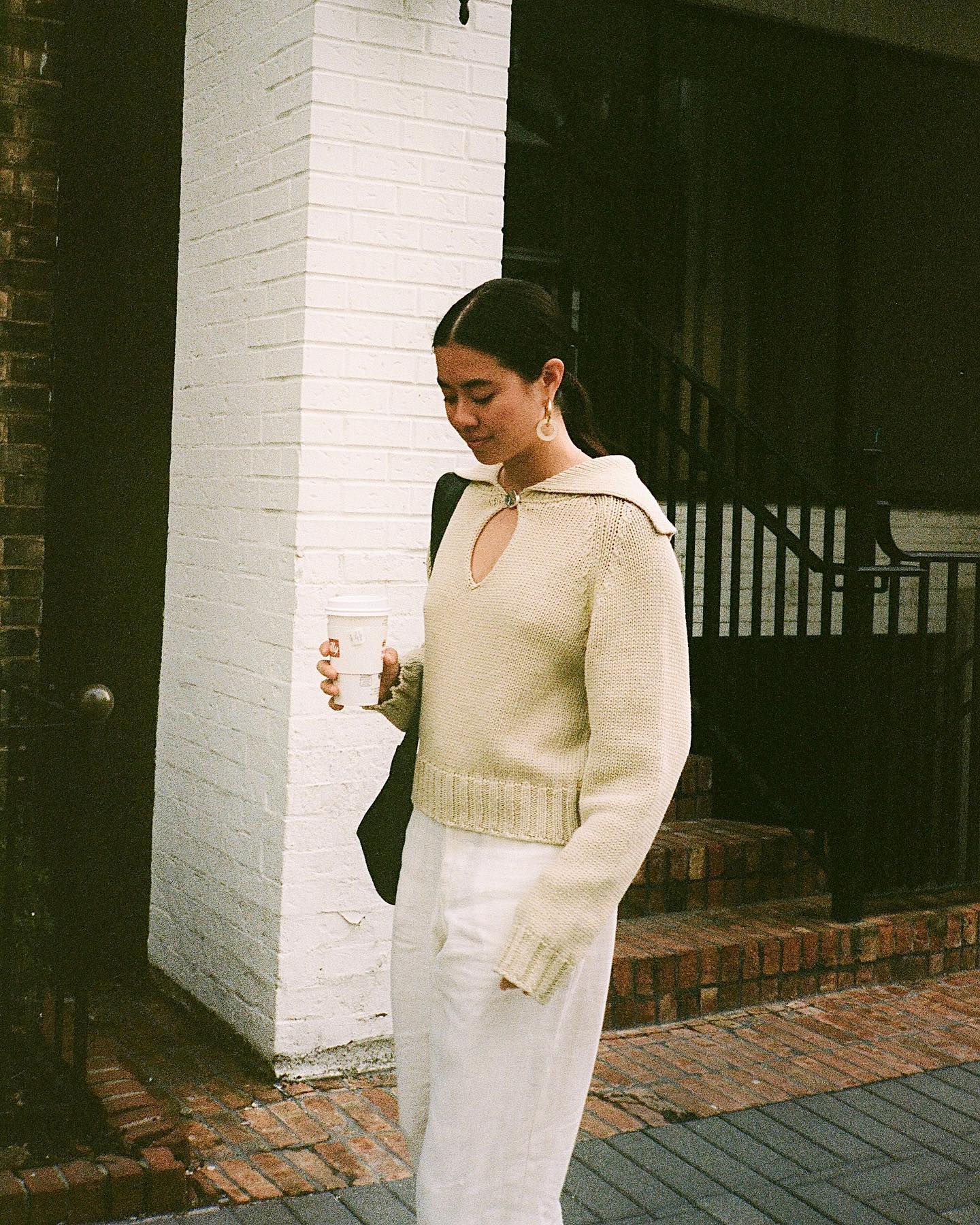 a stylish woman carries a coffee cup while walking on the sidewalk wearing a neutral key-hole sweater, hoop earrings, and white pants
