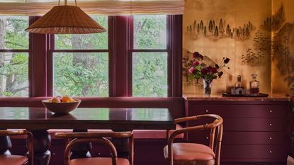 Image of a dining room that has plum purple painted trim and yellow-gold wallpaper. The table is a dark wood, and there is a bench seat on one side and three bamboo chairs with coral cushions on the other.