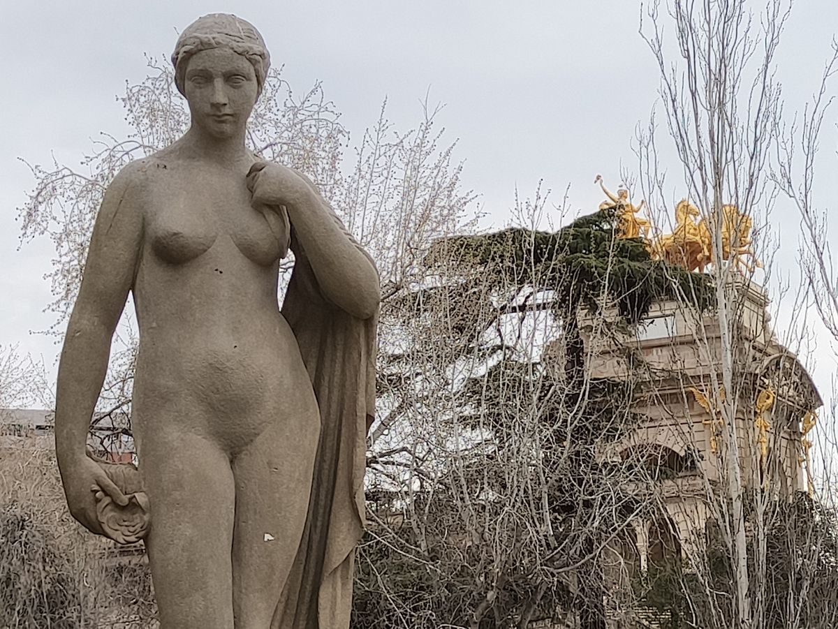 A stone sculpture of a draped woman stands in the foreground, with a large, ornate arch topped by a golden chariot visible through the bare trees in the distance.