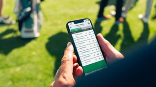 A golfer holds their phone with a live leaderboard on it