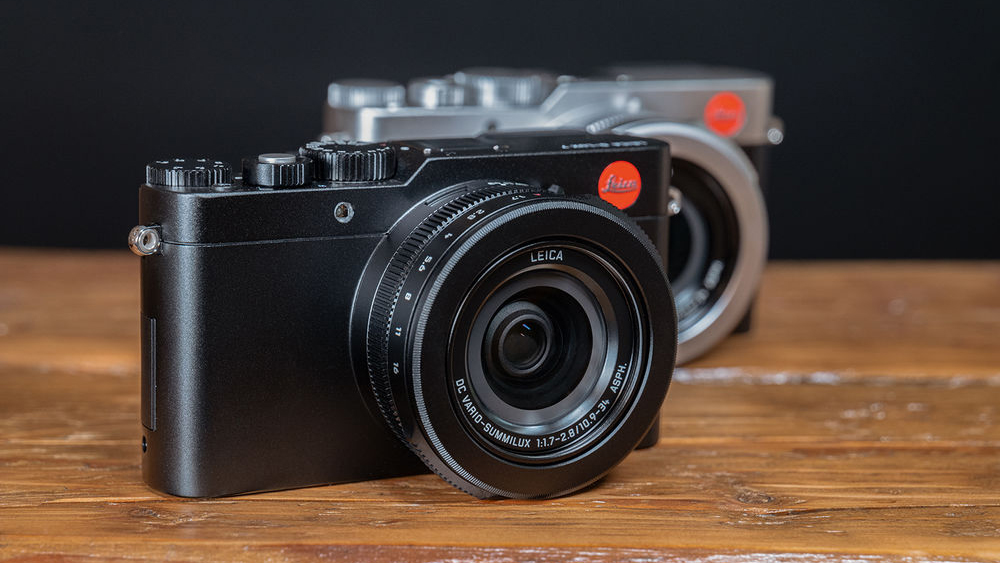 A pair of Leica D-Lux 7 cameras, one in black and one in silver, on top of a wooden surface with a dark background