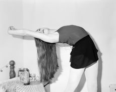 A black and white photo of a young female dancer with long hair bending over backward in front of a table in her room.
