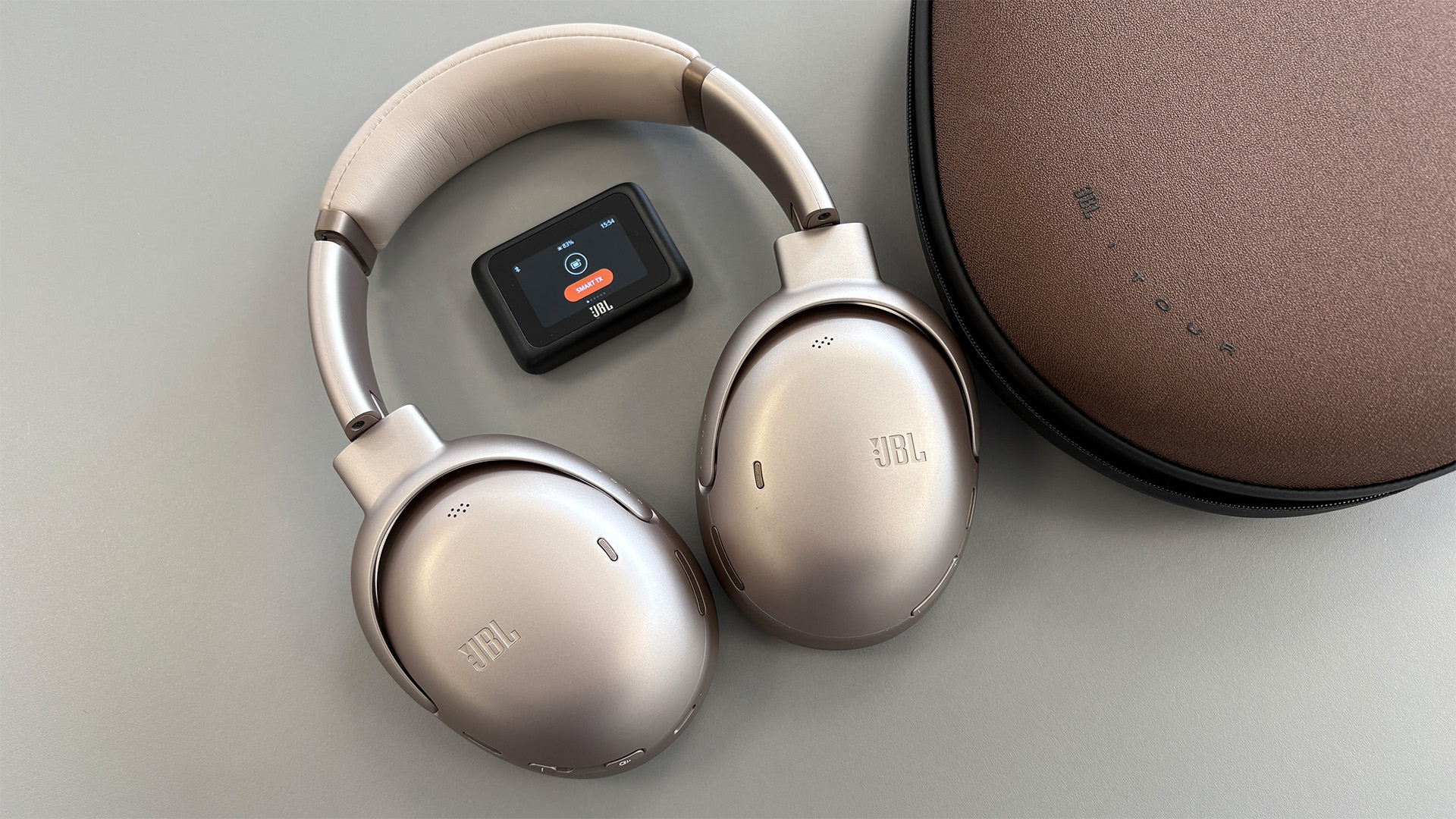 JBL Tour One M3 Smart Tx wireless over-ear headphones on grey surface with Smart Tx transmitter and carry case
