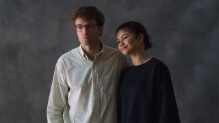 Robert Pattinson in a white button down and Zendaya in a black sweatshirt standing in front of a plain grey background. 