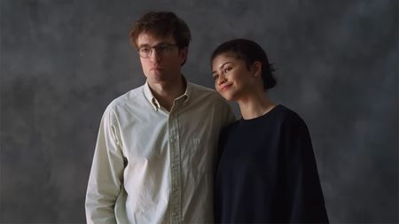 Robert Pattinson in a white button down and Zendaya in a black sweatshirt standing in front of a plain grey background.