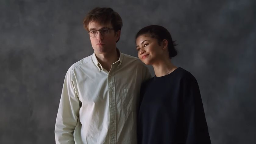 Robert Pattinson in a white button down and Zendaya in a black sweatshirt standing in front of a plain grey background. 