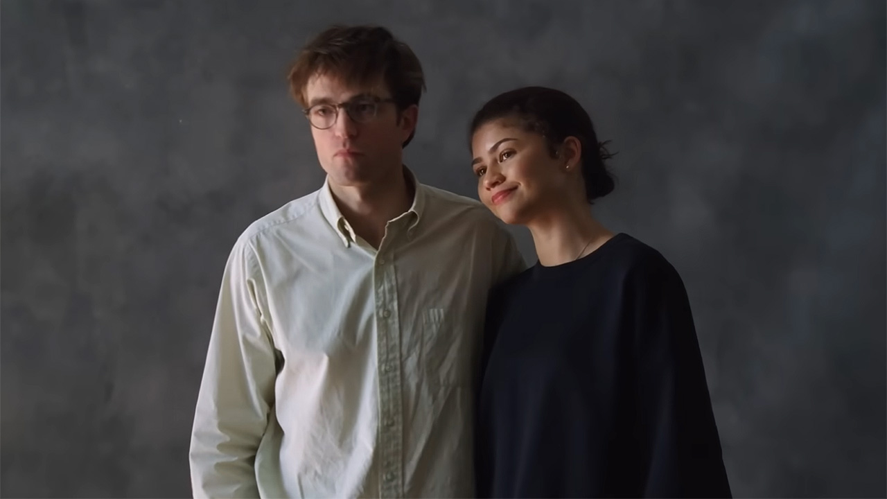 Robert Pattinson in a white button down and Zendaya in a black sweatshirt standing in front of a plain grey background. 