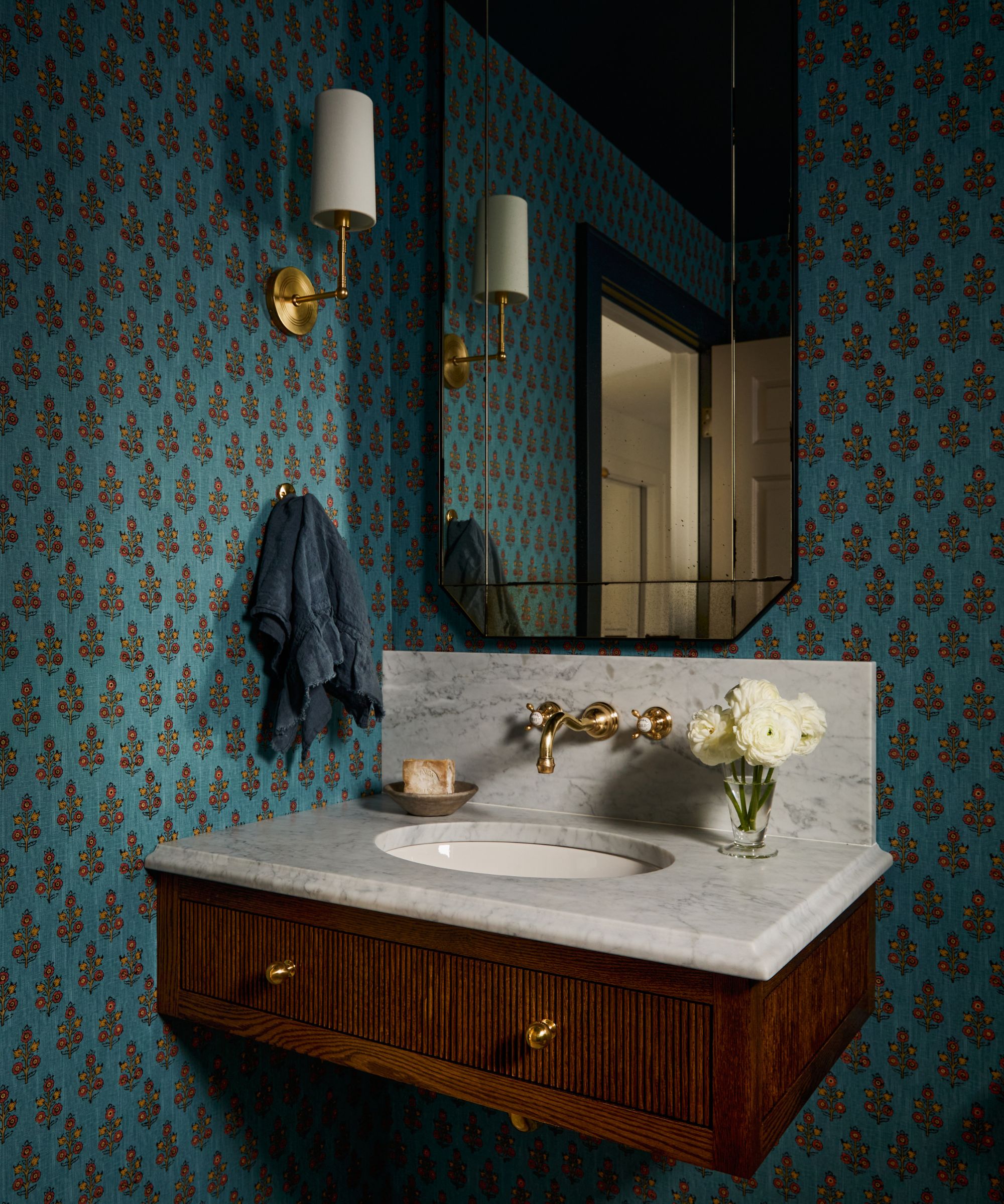 blue bathroom with blue block print floral wallpaper with a small wooden vanity with marble top