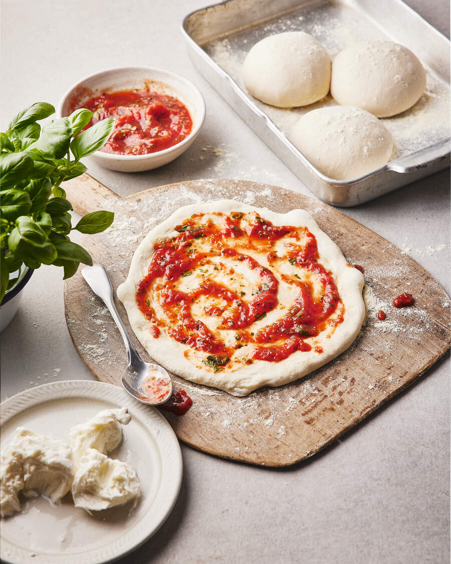 A wooden board with a rolled-out pizza dough topped with tomato sauce and herbs, alongside a bowl of sauce, fresh dough balls, and a plate of creamy cheese.