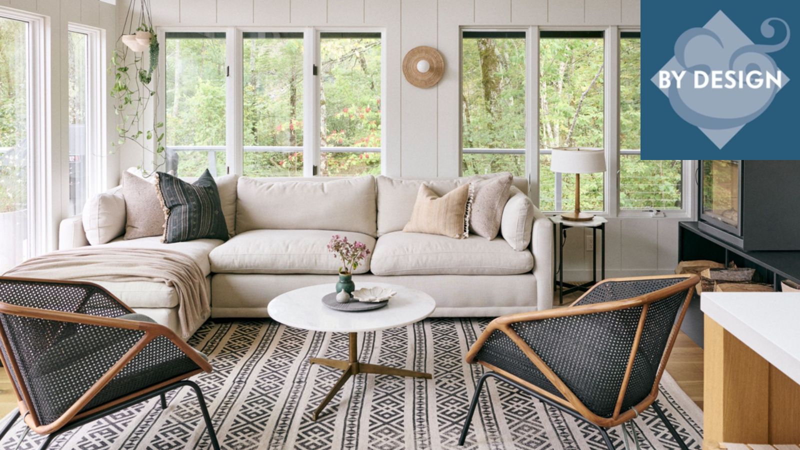 Cozy white living room with panelled white walls, patterned black and white rug, two blac armchairs, cream sofa, fireplace to right of room, large windows with greenery outside