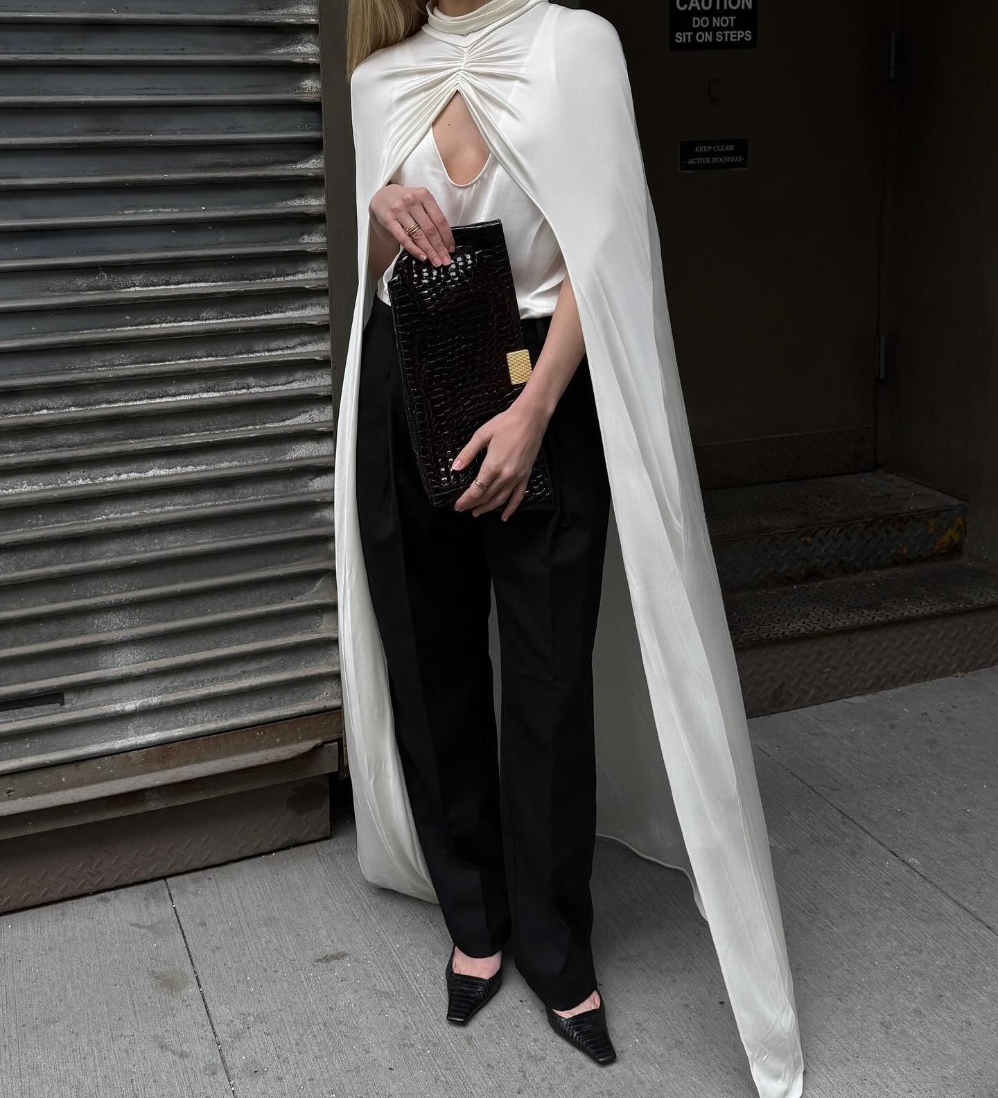 Christie wears a white silk top, white silk cape, black pants, and a black croc clutch.