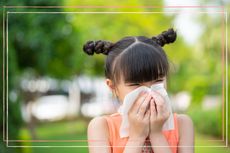 A child sneezing into a tissue whilst standing outside in a green space during summer, suggesting she has hay fever as seasonality is a key difference between hay fever and a cold