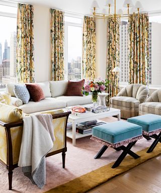 colors that go with light pink, apartment living room with corner aspect, floral yellow drapes, yellow upholstered couch, off white couch, armchairs, coffee table, pair of sky blue stools, pink and yellow rug, retro style pendant