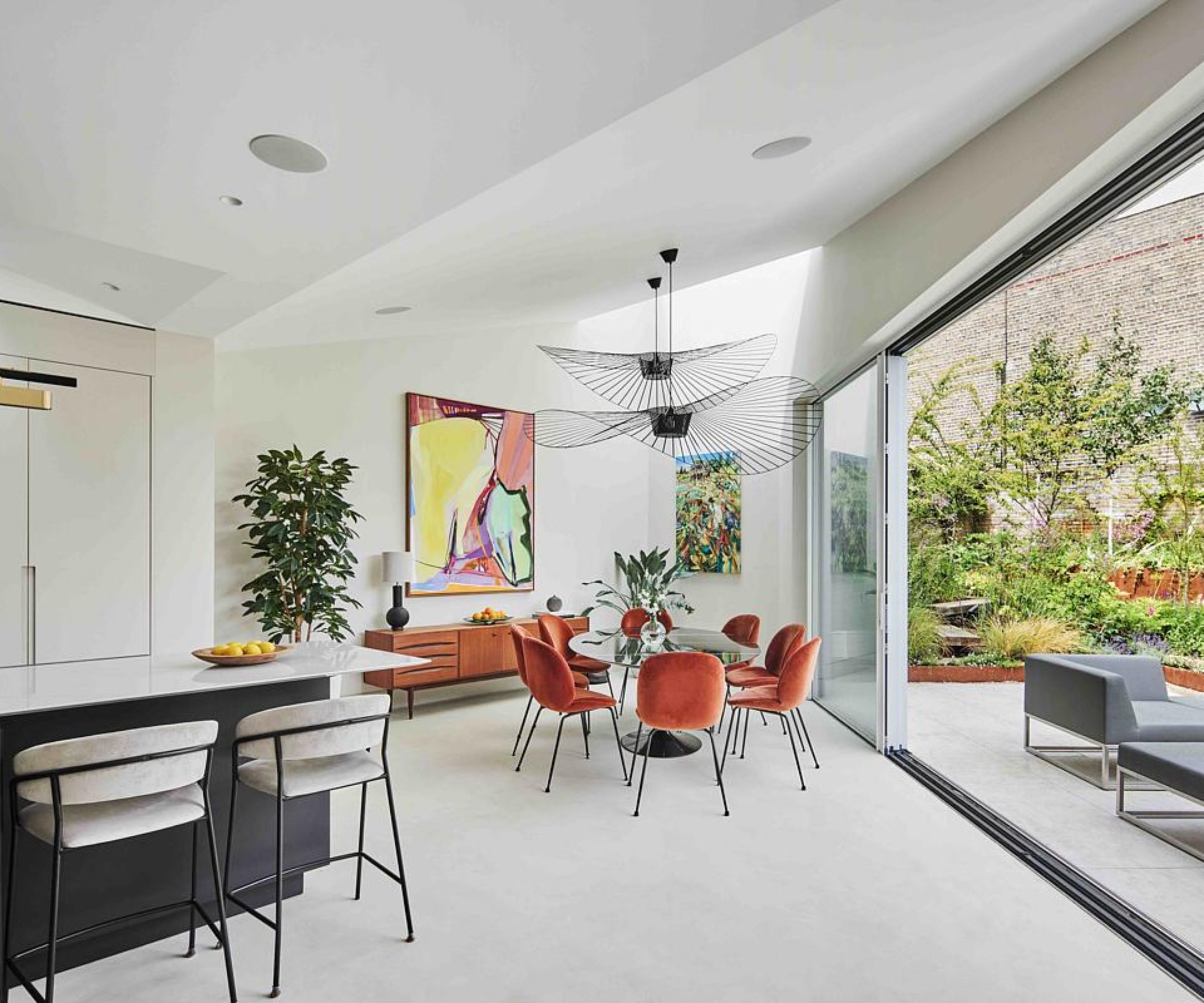 inside single storey extension with angled exterior, large sliding doors, ktichen and angled ceiling levels, dining space to one side with large black pendant lights, orange dining chairs and glass table