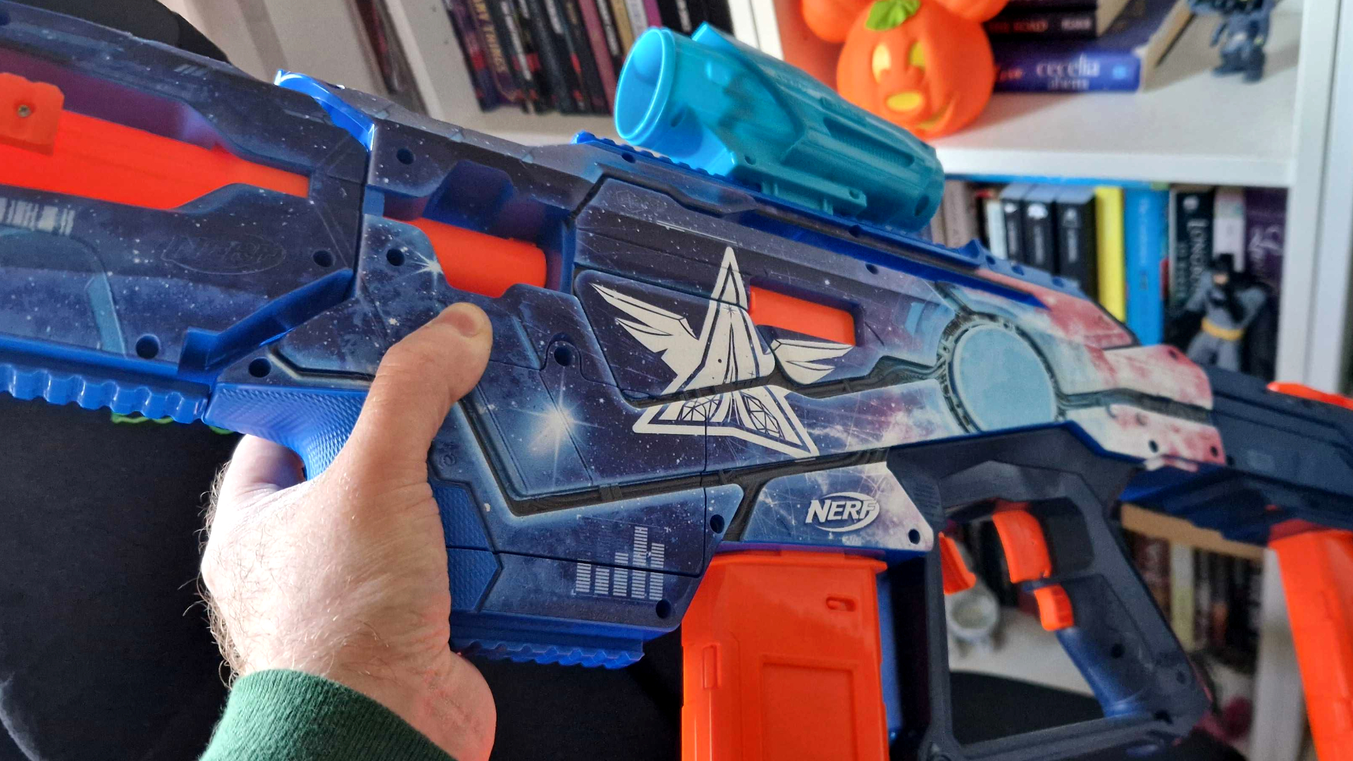 A hand holds the Nerf Loadout Galactic Commander against a bookcase filled with collectibles and books