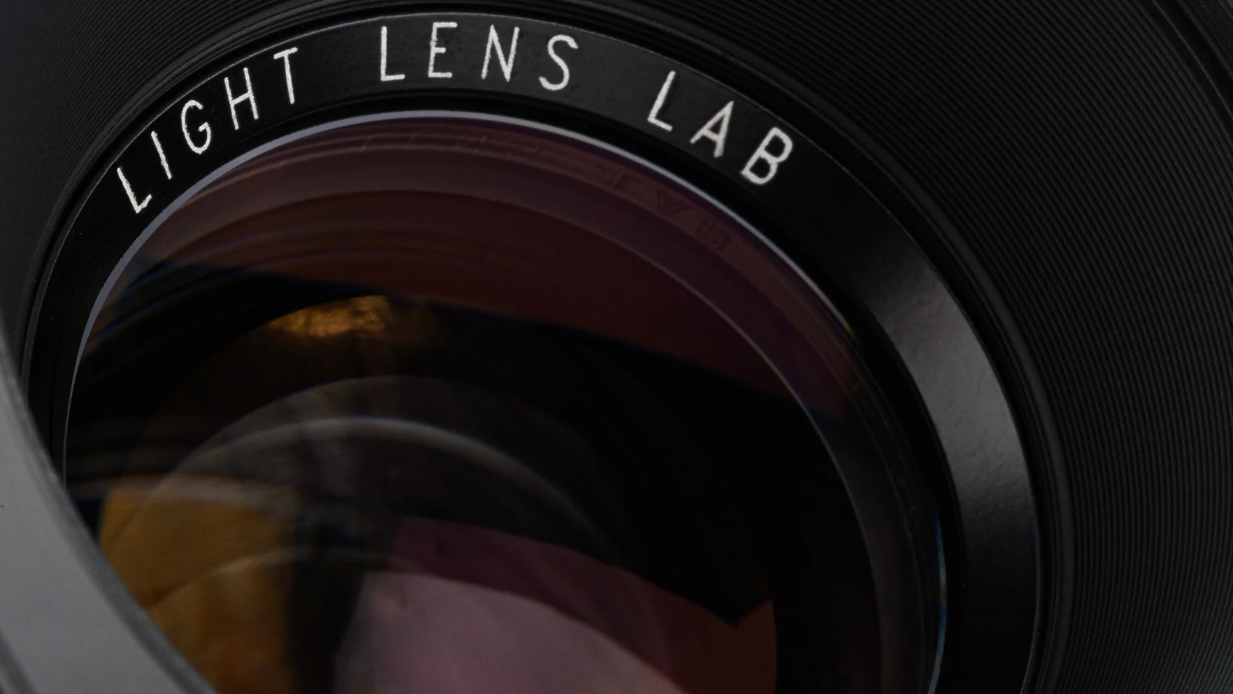 Close-up of a camera lens featuring the engraving "LIGHT LENS LAB" on the black lens surround. The glass reflects warm light