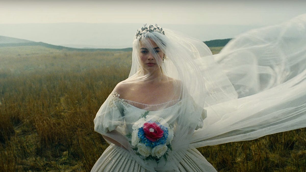 A young blonde woman dressed in a wedding gown stands in a field in front of the camera while holding a colorful, pink, blue, and white bouquet in her hands, wearing a large tiara, and a veil that's been blown by the wind to her left-hand side.
