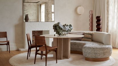 Image of a soft white dining room with a minimalist design scheme. There is a round table with an off-white curved fabric sofa as a bench seat and two wooden dining chairs.