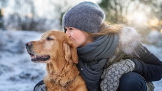 Woman all rugged up in winter clothing outside in the snow kissing the head of her Golden Retriever