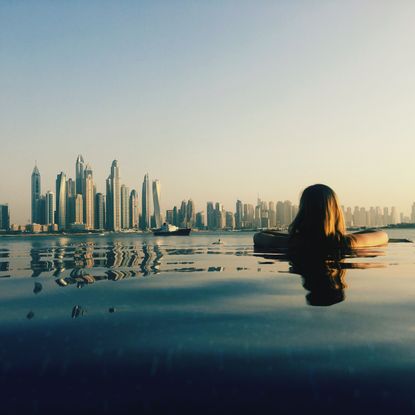 Rear View Of Woman Looking At Modern Building While Swimming In Infinity Pool Against Clear Sky During Sunset