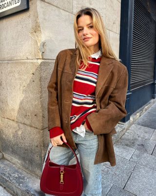 French style influencer Sabina Socol poses on a Paris sidewalk wearing a brown suede jacket, Reformation red striped sweater, light-wash jeans, and a red Gucci Jackie bag.