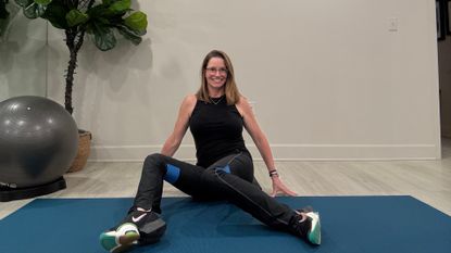 An employee at Femina Physical Therapy performs a hip mobility move on an exercise mat. They are sat with their hands behind their body, legs in front, feet on the floor and knees bent. They are dropping their knees to the right side and smiling. Behind them there is a plant and exercise ball. 