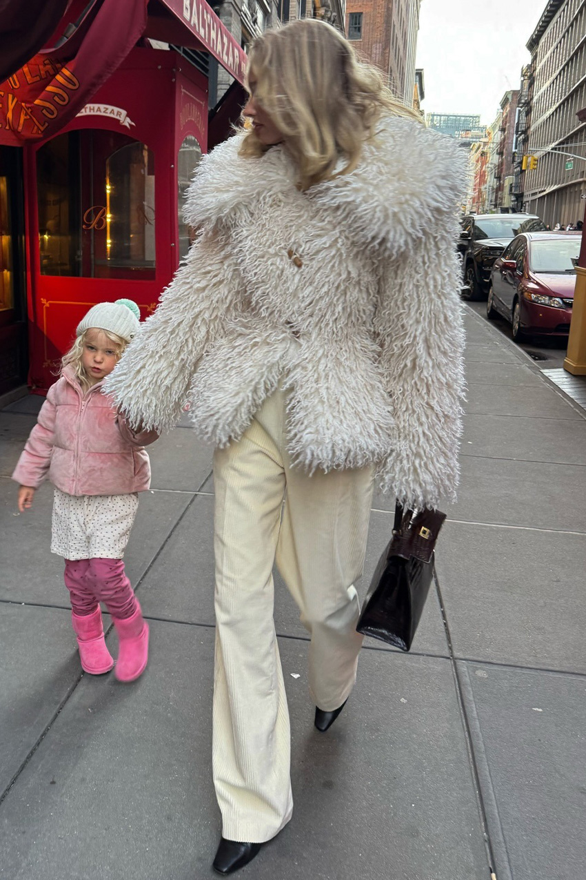 A woman on the sidewalk in New York wearing a white shaggy fur coat with ivory wide leg pants, black ankle boots, and a black structured handbag.