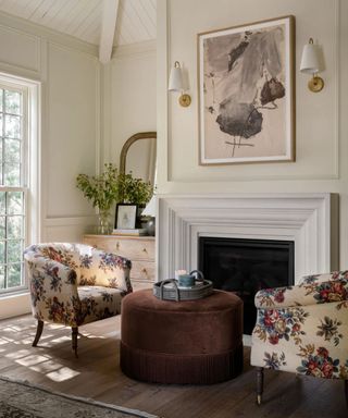 small seating area beside a fireplace with a velvet fringed ottoman and two antique style floral armchairs