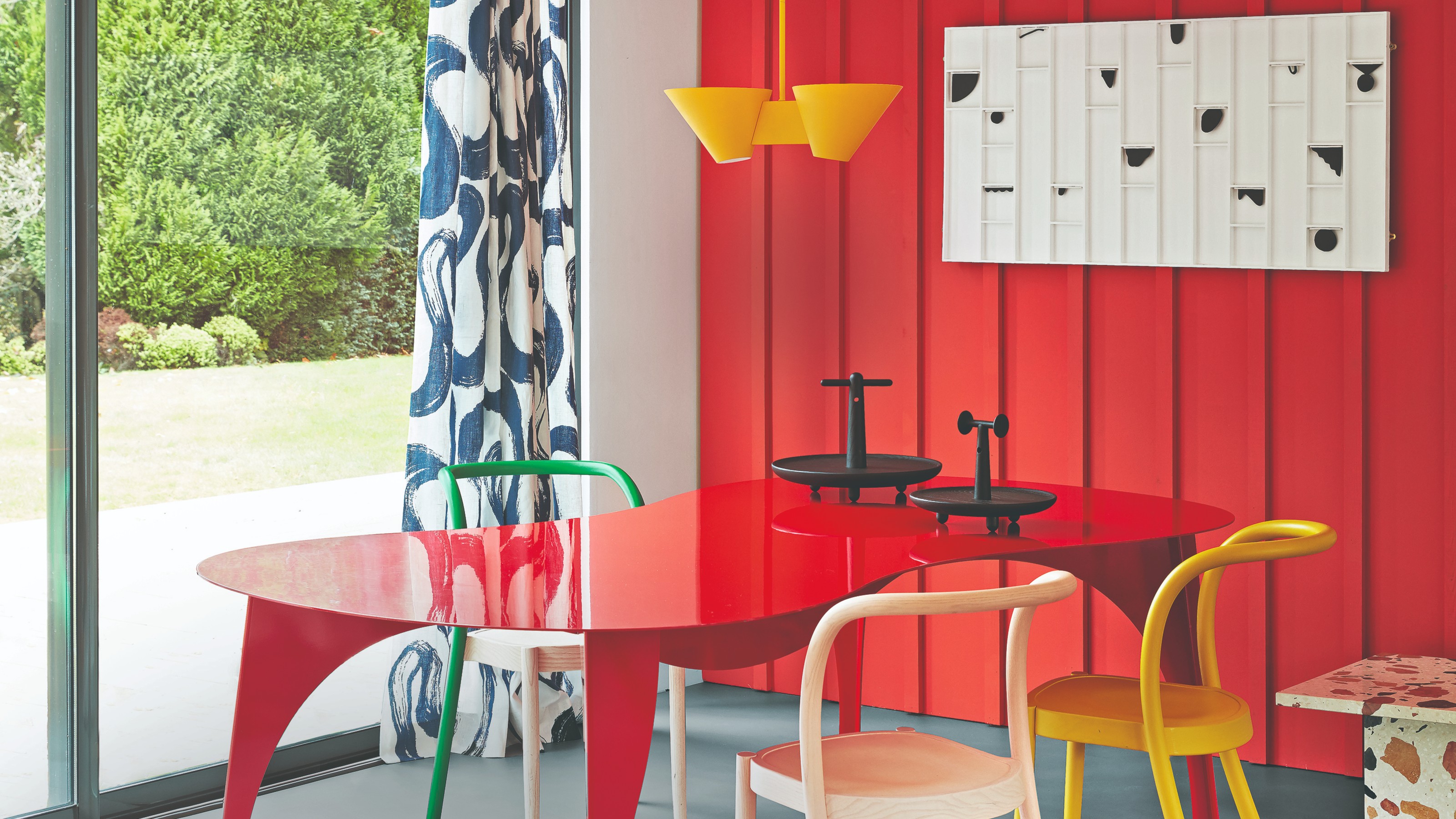 A dining room with a panelled red feature wall, a red organically shaped dining table and a yellow dining chair and pendant light
