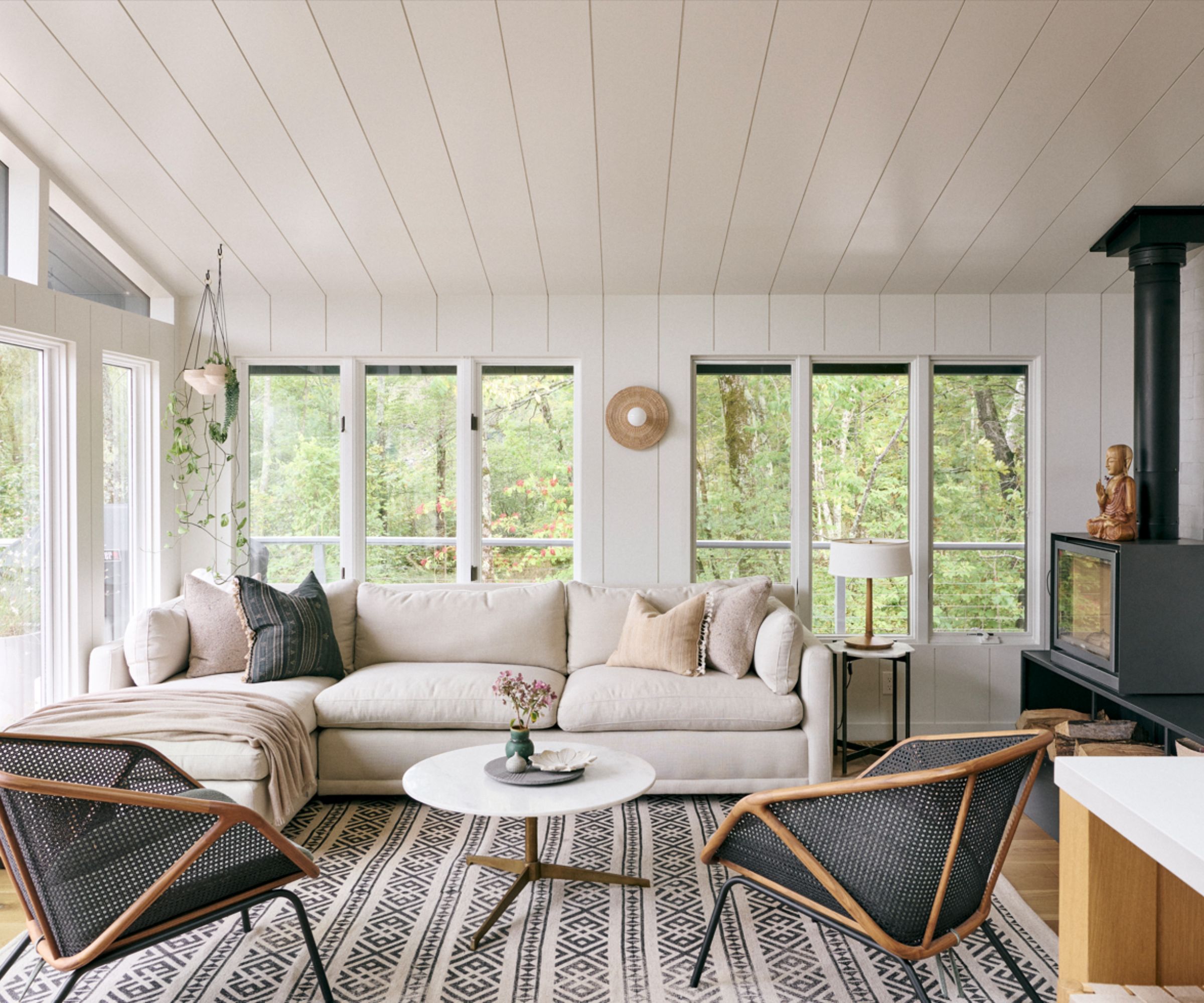 Cozy white living room with panelled white walls, patterned black and white rug, two blac armchairs, cream sofa, fireplace to right of room, large windows with greenery outside