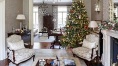 A festive living room with a large lit Christmas tree to the right. In the foreground is a large square ottoman on top of a cream rug with matching cream upholstered chairs with wooden legs. To the right is a white mantelpiece with a floral garland. A room is seen beyond with a matching cream carpet and dark wood furniture, and two windows with white patterned curtains.