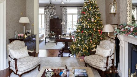 A festive living room with a large lit Christmas tree to the right. In the foreground is a large square ottoman on top of a cream rug with matching cream upholstered chairs with wooden legs. To the right is a white mantelpiece with a floral garland. A room is seen beyond with a matching cream carpet and dark wood furniture, and two windows with white patterned curtains.
