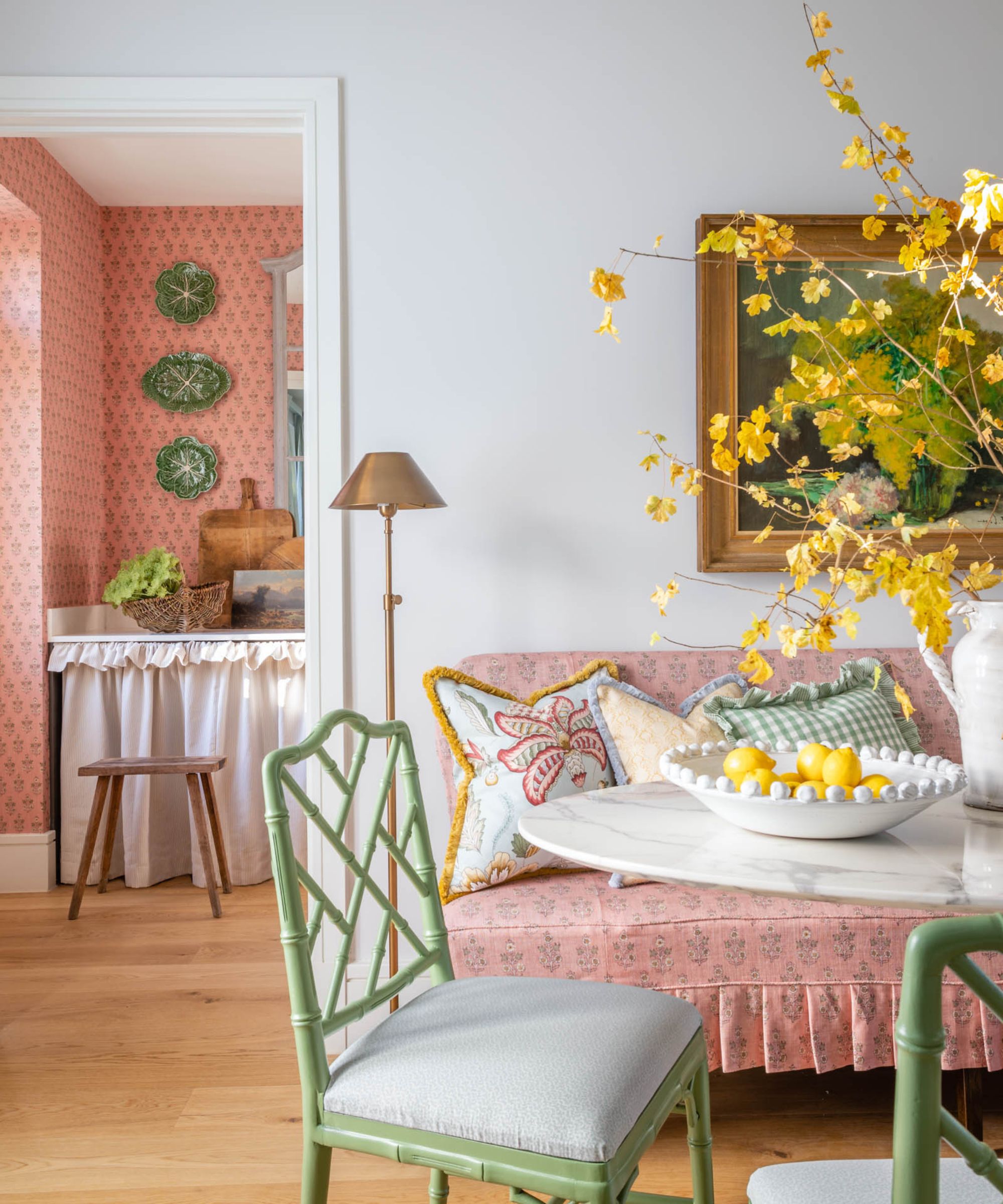 a breakfast nook with a pink floral banquette seat, marble table, green bamboo dining chairs and a view into a pink floral wallpapered kitchen with cabbage plates and a white cabinet skirt