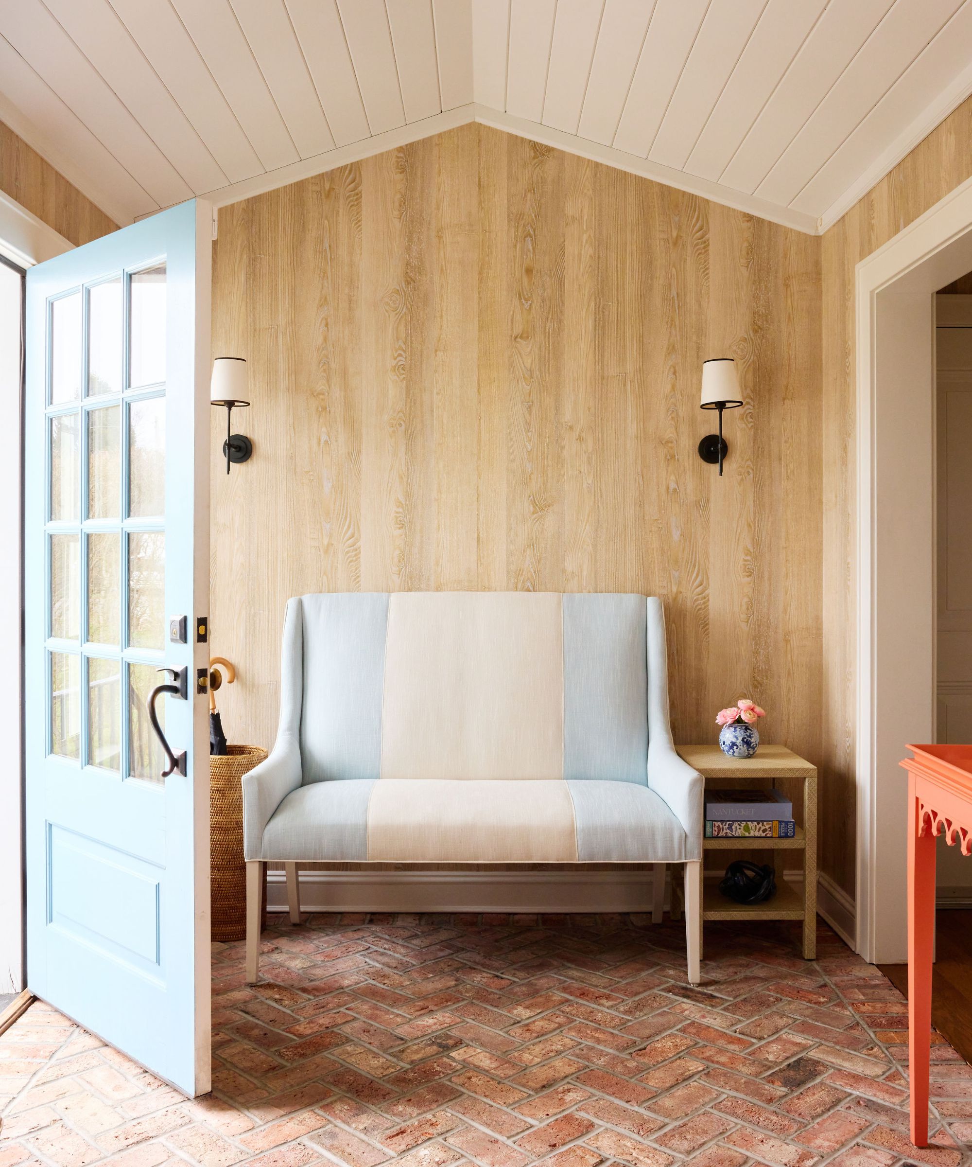 Entryway with wood panelled walls, an exposed brick floor, a pale blue and cream striped loveseat, an orange table, a pale blue front door and a shiplap white ceiling.