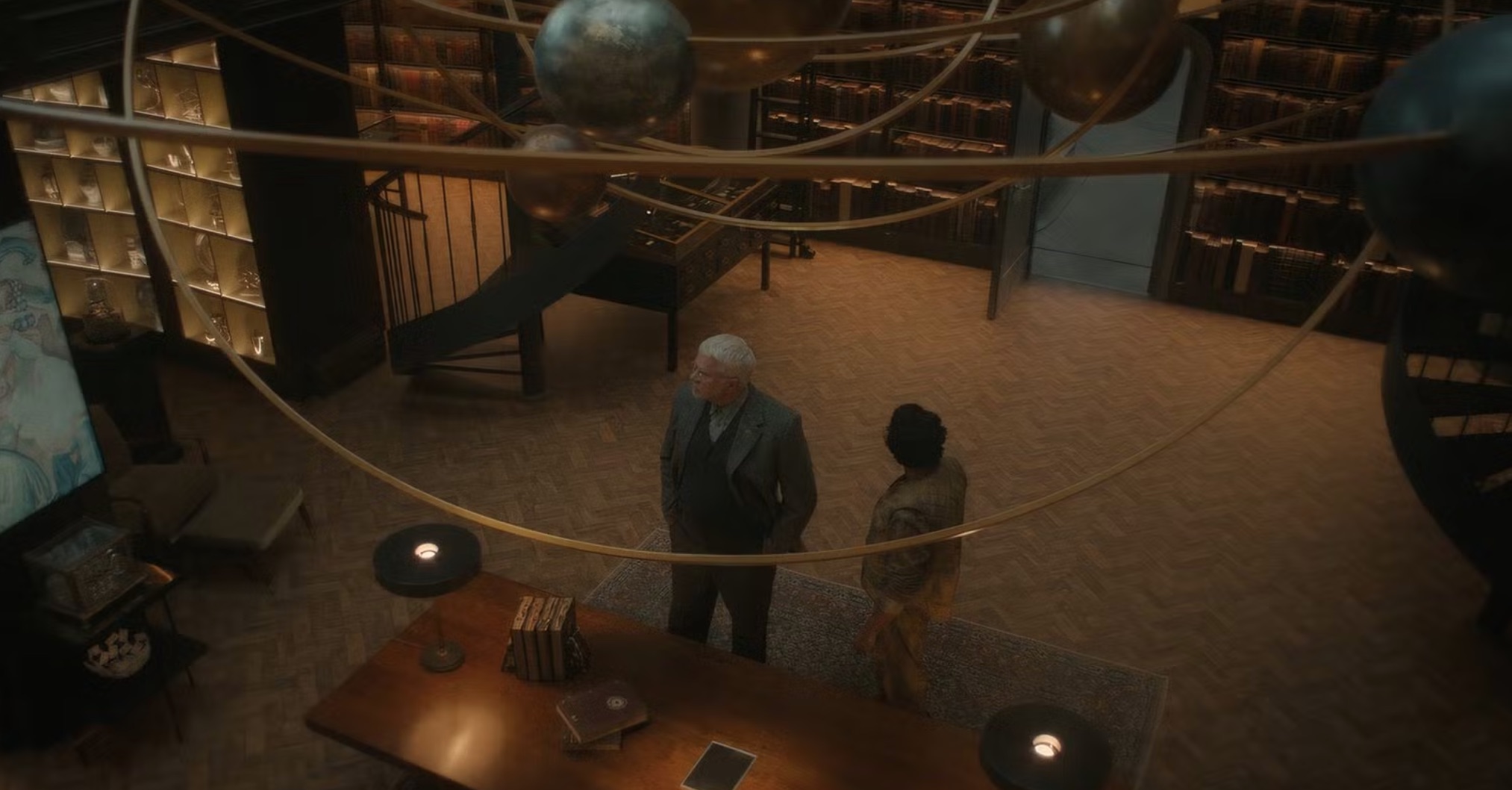 Two men inside a library-like vault with a solar system model overhead