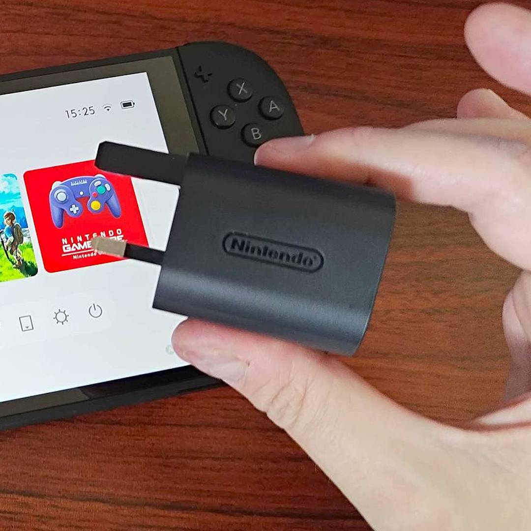 Hand holding Nintendo Switch 2 AC adapter with console in backdrop on desk.