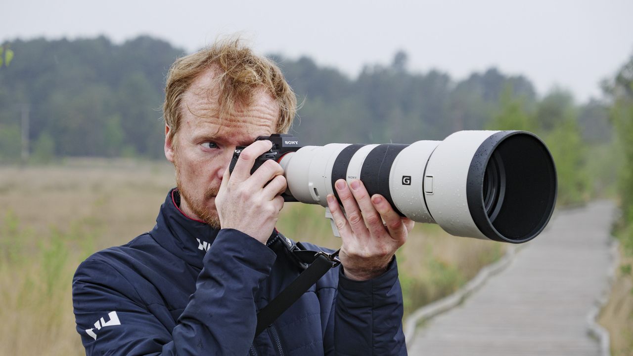 Man holding the Sony A7R VI up to his eye with the FE 400-800mm F6.3-8 G OSS supertelephoto zoom lens attached, he's standing on a boardwalk in a nature reserve