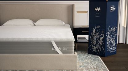 AirFoam&reg; Luxe Mattress Topper on a beige upholstered bed with two white pillows. Beneath the bed is a patterned rug and dark wooden flooring, and to the right is a large dark blue box.