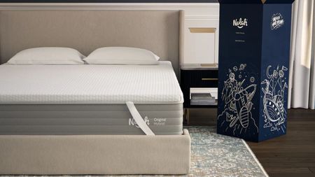AirFoam® Luxe Mattress Topper on a beige upholstered bed with two white pillows. Beneath the bed is a patterned rug and dark wooden flooring, and to the right is a large dark blue box.