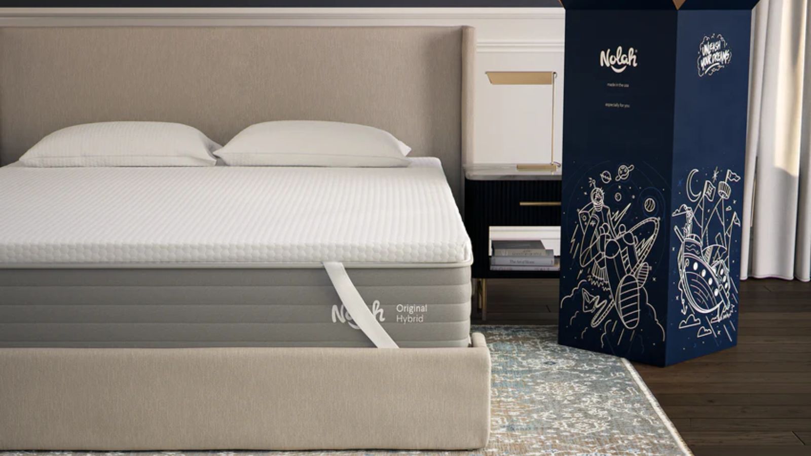 AirFoam&reg; Luxe Mattress Topper on a beige upholstered bed with two white pillows. Beneath the bed is a patterned rug and dark wooden flooring, and to the right is a large dark blue box.
