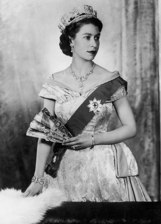 Studio portrait of Queen Elizabeth II holding a fan while wearing a brocade dress.