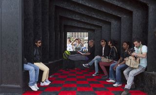 Interior view of a black and red structure with checked flooring. There are several people sitting inside and two people can be seen outside through a square opening at the back