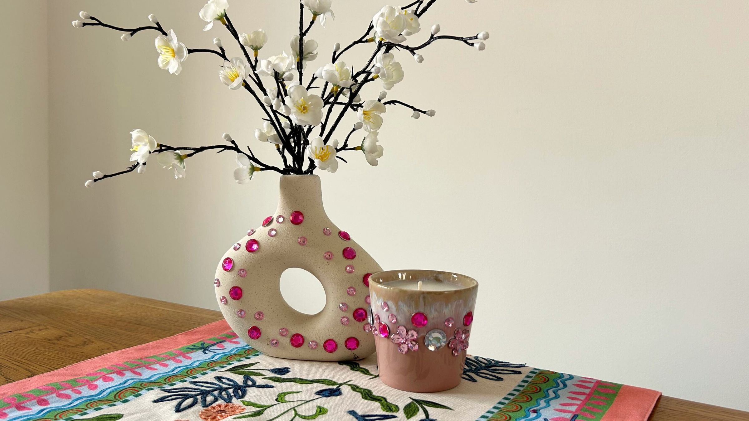 A white vase bedazzled in pink gems next to a pink bedazzled candle on a table in front of a white wall