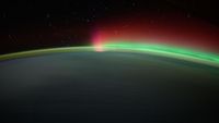 a wash of green aurora, crowned in red, wisps over the top of a night-shadowed earth.