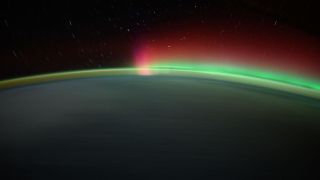 a wash of green aurora, crowned in red, wisps over the top of a night-shadowed earth.