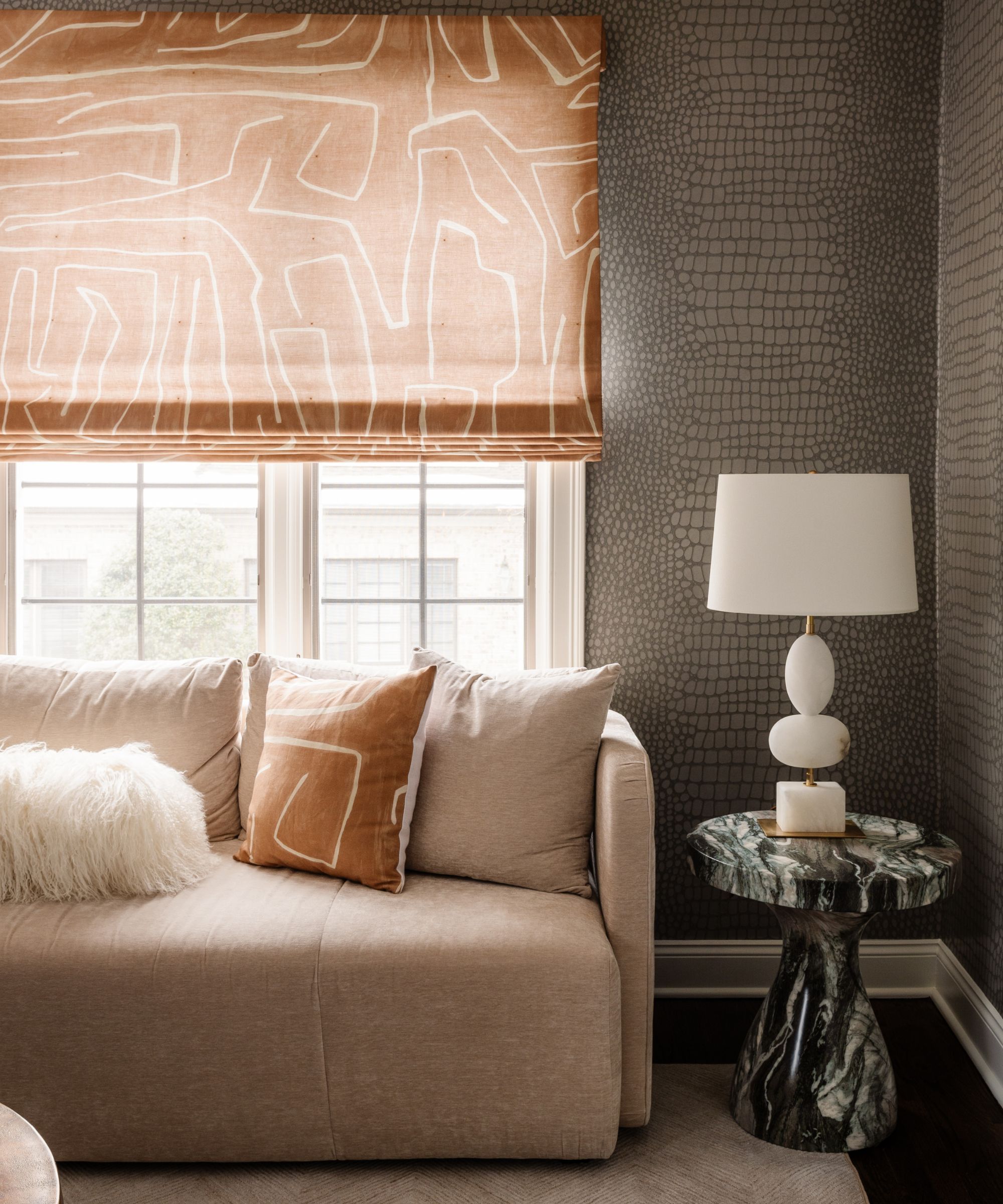 Living room with snakeskin wallpaper on the walls and a beige couch, marble side table with white lamp and a patterned orange blind on a window above a sofa