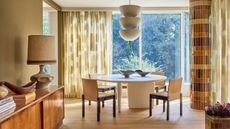 modern dining room with patterned sheer curtains, a round white dining table with leather chairs and a pendant hanging overhead, a structural column that's been tiled, timber floors, a timber console with lamp, books, and decor