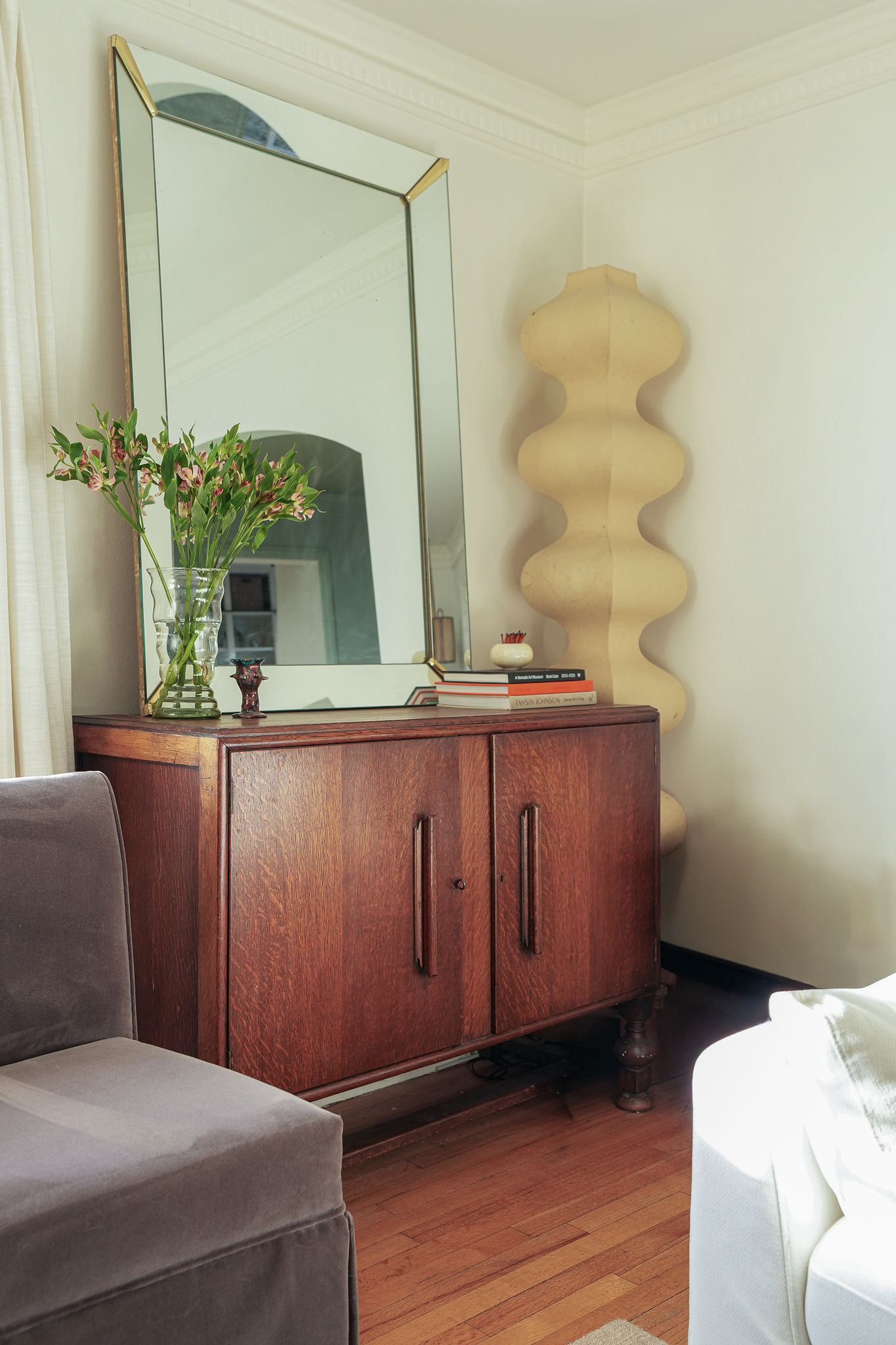 The corner of a white living room with wooden floors. There is a wooden console table with a mirror on it, a vase, a stack of books, and a paper lantern floor lamp in the corner.